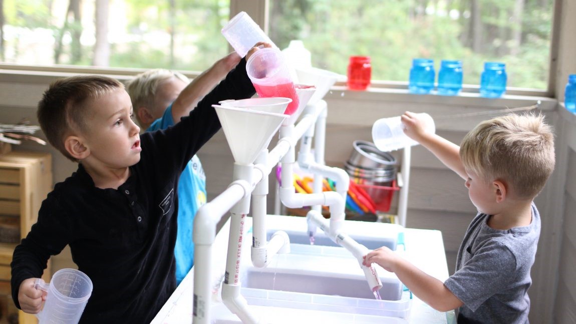 Preschool boys in a science class pour water into different funnels connected to a network of PVC pipes