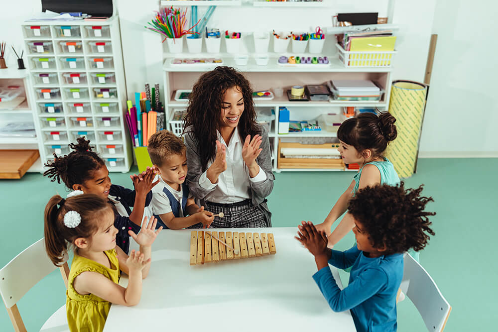 A female teacher at a table with preschool kids all clapping their hands