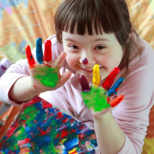 Young white girl with down syndrome holds up paint covered hands and smiles at camera. She also has a dot of paint on her nose.