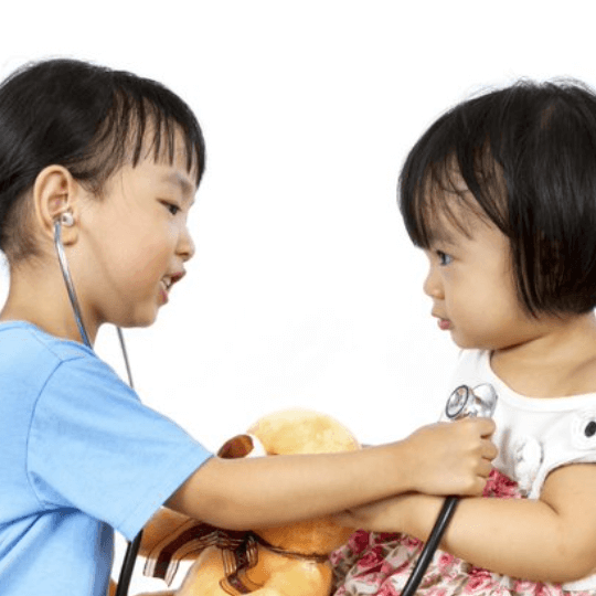 Two children sit face to face, the child on the left wears a stethoscope and holds it up to the chest of the child on the right. She is holding a stuffed bear. Both children appear to be Asian girls.