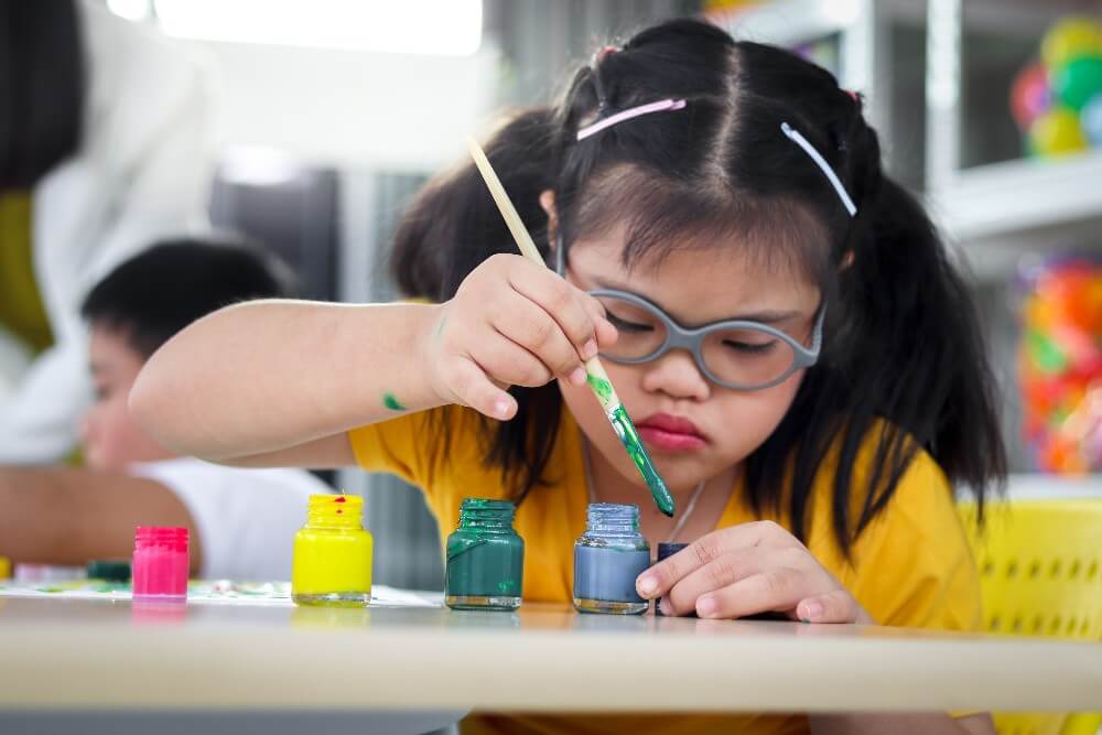 A young Asian child with pigtails and barrettes, wearing glasses, dips a paint brush in pots of paint.