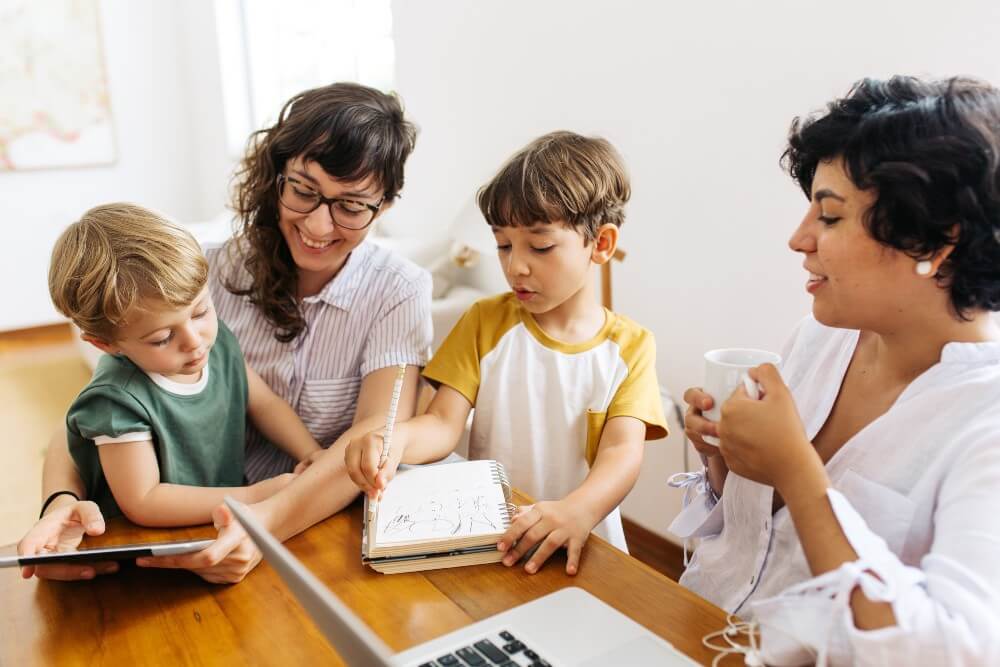 Image shows a mixed-race family with two moms and two children interacting at a table with a tablet, notebook, and laptop.