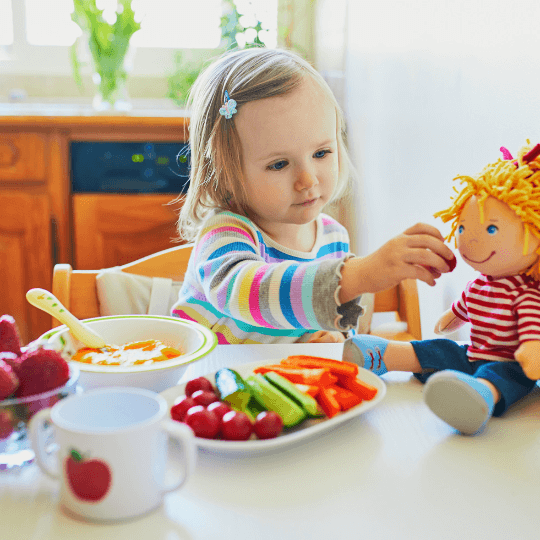 A white child with blond hair sits before a table with a plate of cut veggies, a bowl of food, a mug, and a container of strawberries. Also on the table is a white doll with yellow hair. The child is pretending to feed a strawberry to the doll.