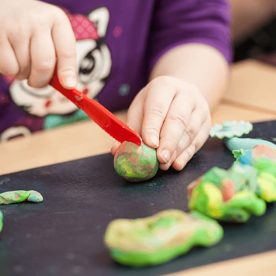 An image of homemade, multicolor play dough sits in the foreground. A white child's hands hold a red plastic knife and cuts one of the pieces of play doh.