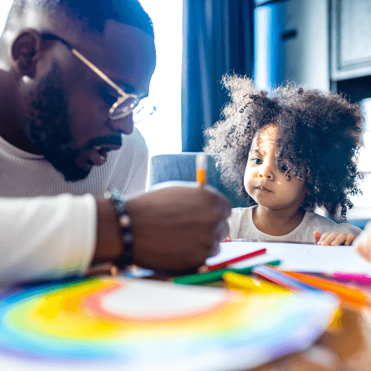 Black father in foreground holds marker and colors on a sheet of paper on a table. Also seated at the table is a young Black or mixed race child watching him draw. The father is wearing glasses, an earring, and a beaded bracelet. The child has curly natural hair.